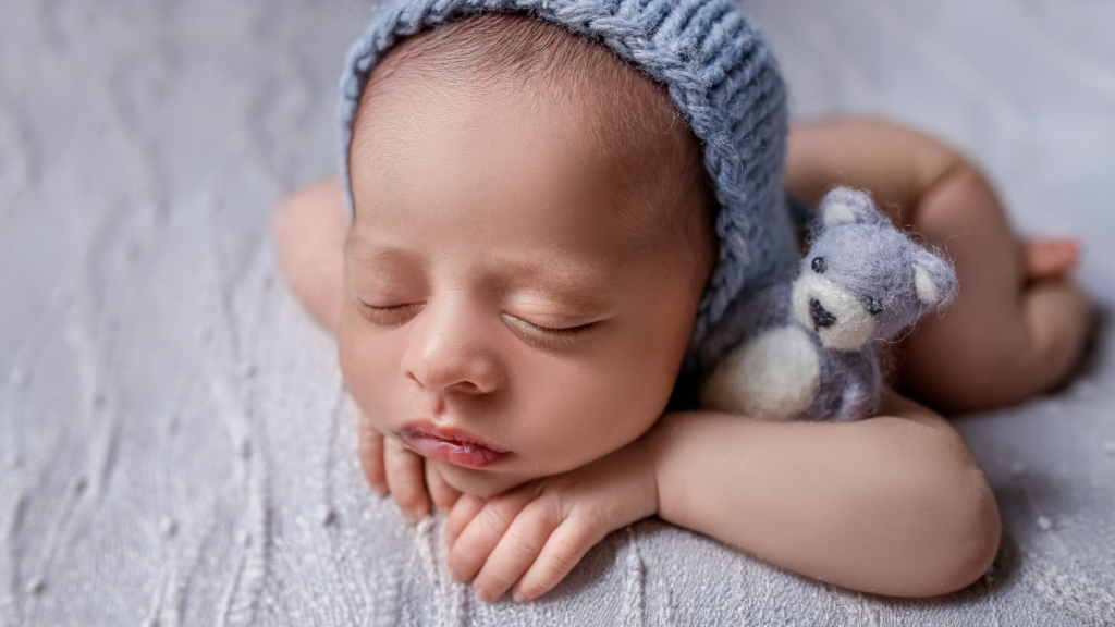 Foto de um recém-nascido dormindo com um gorro azul e segurando um ursinho de pelúcia, após a pós-produção fotográfica de carla toscano pós-produção fotográfica, com pele suavizada e tonalidade aprimorada